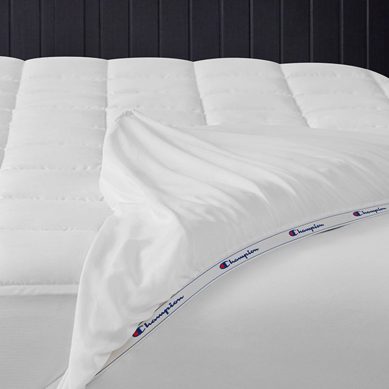 A neatly made bed with a white comforter and sheets, featuring a blue and red Champion logo band along the edge of the top sheet, is complemented by the Champion Ultimate Protection Mattress Pad for added water-resistant protection.