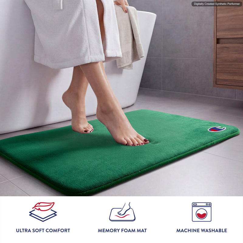A person stands barefoot on a green Champion Logo Patch Memory Foam Bath Mat in a modern bathroom, with only their lower legs visible. Icons below emphasize the ultra-soft comfort, memory foam support, and machine-washable, skid-resistant features of this Champion bath mat.