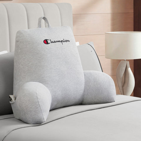 A Champion Jersey Backrest Pillow with armrests in gray rests on a neatly made bed with matching gray bedding. In the background, a white lamp and part of a wooden headboard can be seen.