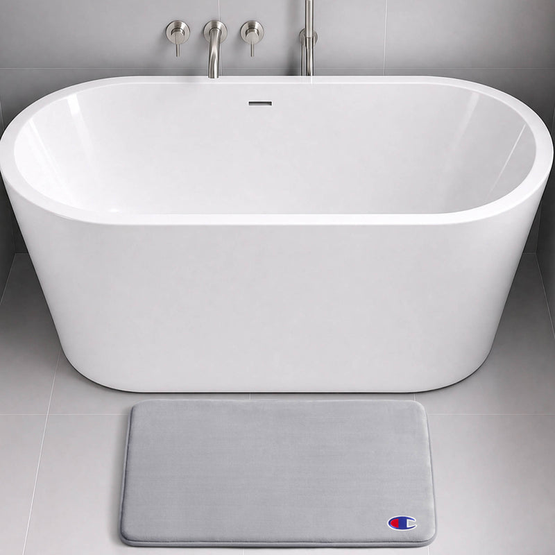 Modern white freestanding bathtub with chrome fixtures, set against gray tiled walls and floor. In front of the tub lies a light gray, skid-resistant Logo Patch Memory Foam Bath Mat from Champion, featuring a small Champion logo.