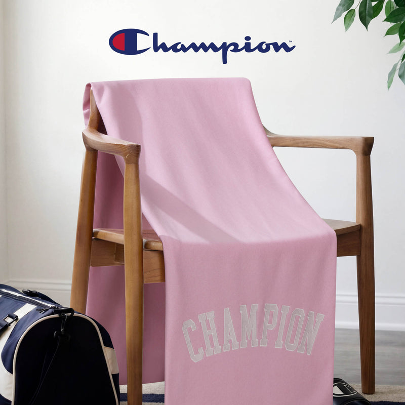 A pink Jersey with Fleece Backing Throw Blanket by Champion, made from soft jersey knit, is draped over a wooden chair, with the Champion logo displayed on the wall above. A navy and white duffel bag sits on the floor beside the chair.