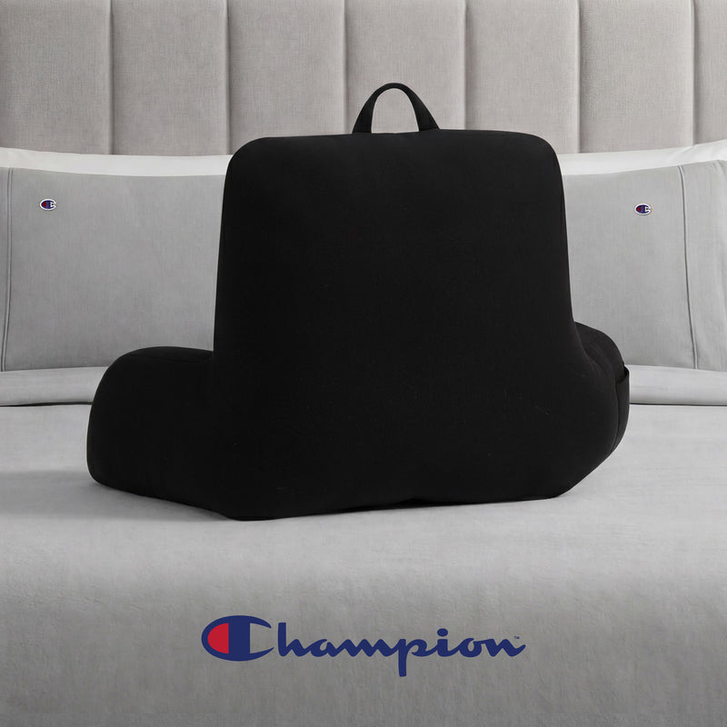 A Champion Jersey Backrest Pillow in black, featuring armrests, is placed on a bed with gray Champion logo bedding. The Champion logo appears at the bottom center of the image.