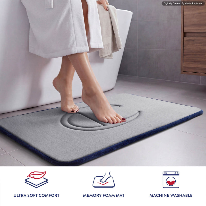A person steps onto a gray Quilted Logo Memory Foam Bath Mat by Champion in a bathroom. Icons and text below highlight its ultra soft comfort, memory foam construction, and machine washable convenience.