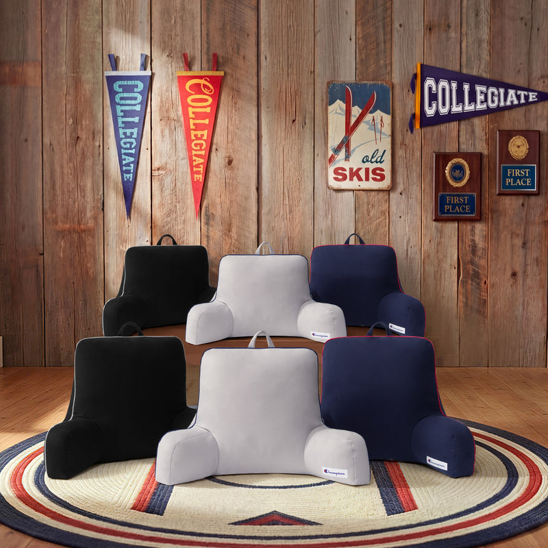 Six Champion Faux Wool Backrest Pillows with supportive arms, in black, gray, and navy, are arranged on a circular rug in a wood-paneled room adorned with collegiate decor, wall signs, and first place plaques. Each pillow is crafted from soft faux wool for enhanced comfort and style.