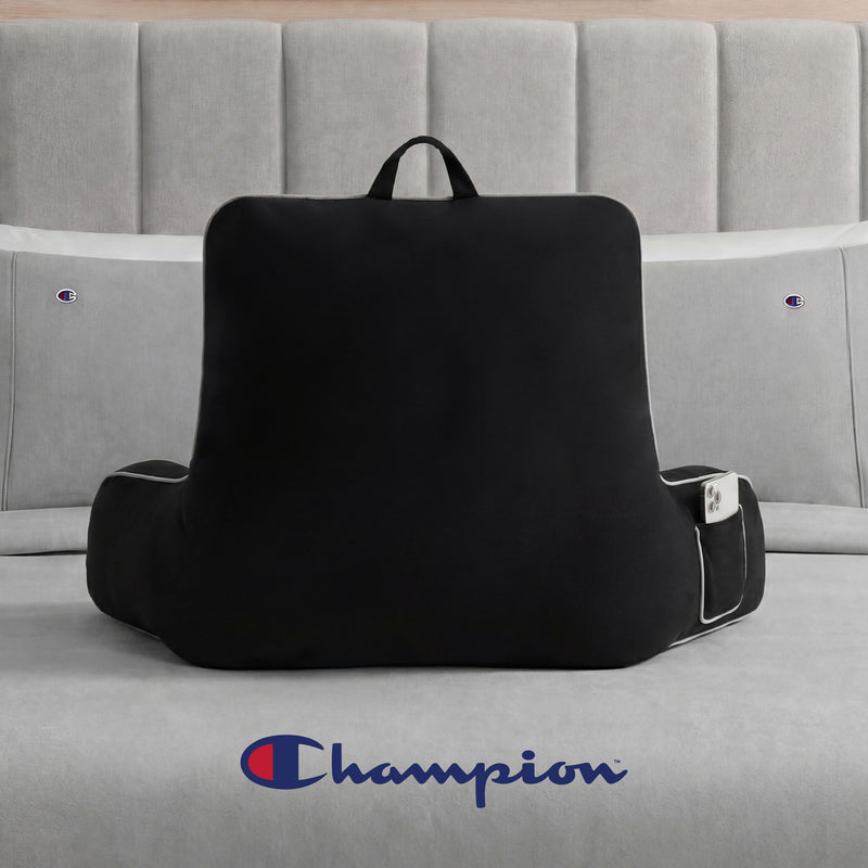A Champion Faux Wool Backrest Pillow in black, featuring supportive arms and crafted from cozy faux wool for comfort and style, rests on a gray bed adorned with Champion logo bedding, while the iconic Champion logo is displayed on the wall above.