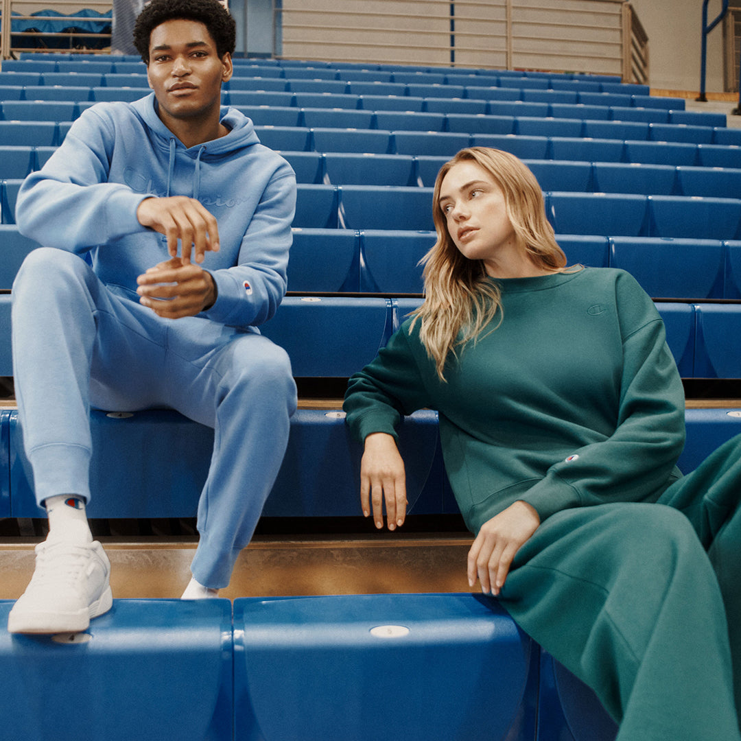 Two models wearing matching loungewear sets, one in light blue and one in dark green, sitting on blue stadium seating in an indoor arena.