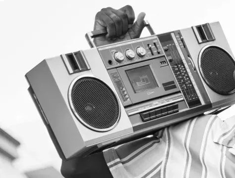 A person holds a large vintage boombox on their shoulder; the image is in black and white and part of the persons arm and striped shirt are visible.