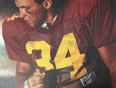 A football player wearing a maroon jersey with the number 34 in large yellow print stands with his head turned to the side, showing eye black on his face.