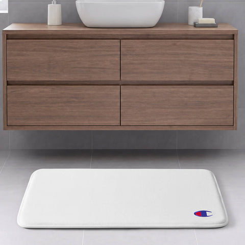A white Champion Logo Patch Memory Foam Bath Mat with a skid-resistant backing and the iconic logo rests on a gray tile floor in front of a floating wooden bathroom vanity featuring four drawers and a white vessel sink.