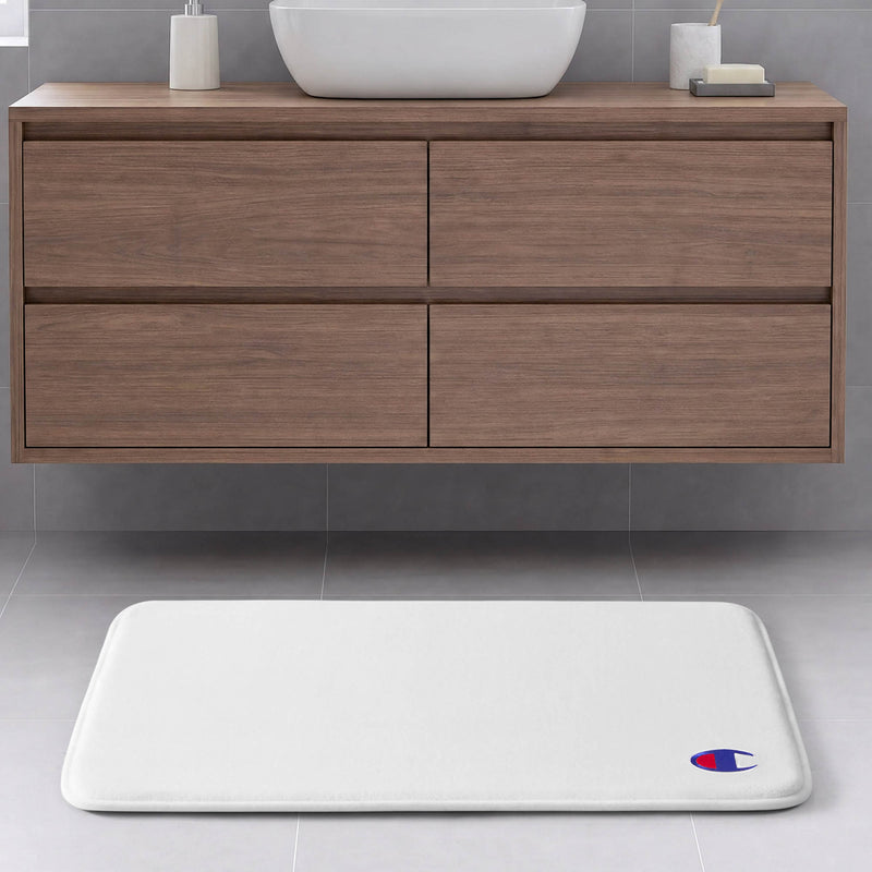 A white Champion Logo Patch Memory Foam Bath Mat with a skid-resistant backing and the iconic logo rests on a gray tile floor in front of a floating wooden bathroom vanity featuring four drawers and a white vessel sink.