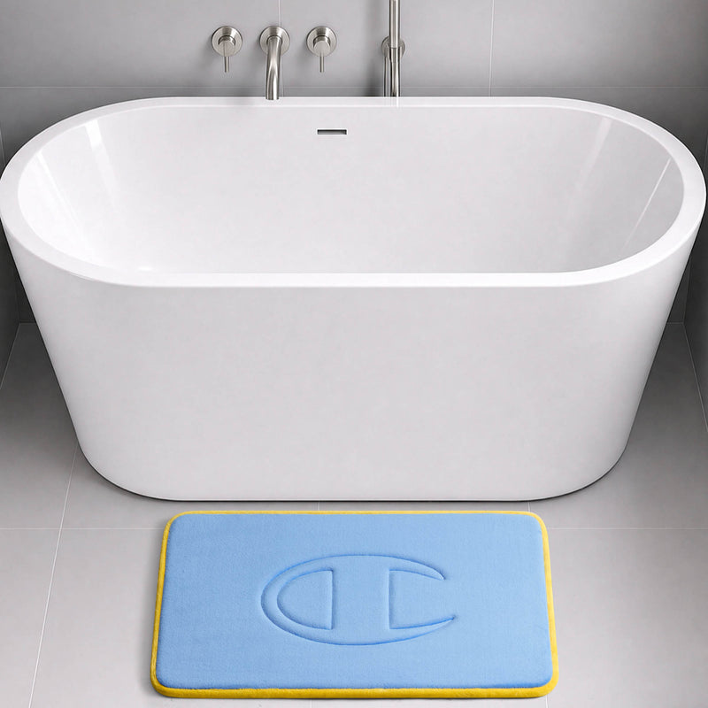 A modern white bathtub with wall-mounted faucets is set against a gray tiled wall. In front of the tub is a rectangular blue Quilted Logo Memory Foam Bath Mat from Champion, featuring a yellow border and raised design, bringing both style and comfort to your bathroom.