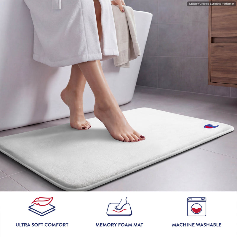 A person in a white bathrobe steps onto a Champion Logo Patch Memory Foam Bath Mat in a modern bathroom. Icons below highlight comfort, memory foam support, skid-resistant design, and the mat’s machine washable convenience.