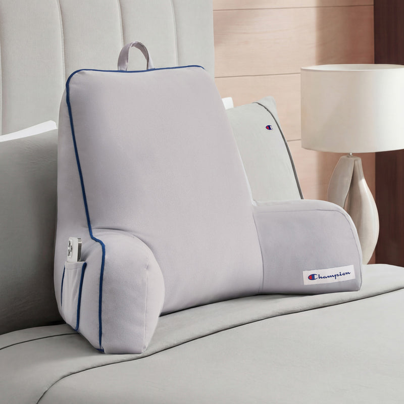 A Champion Faux Wool Backrest Pillow in light gray, featuring supportive arms, blue piping, and convenient side pockets, sits upright on a neatly made bed with gray bedding beside a white lamp on a wooden nightstand.