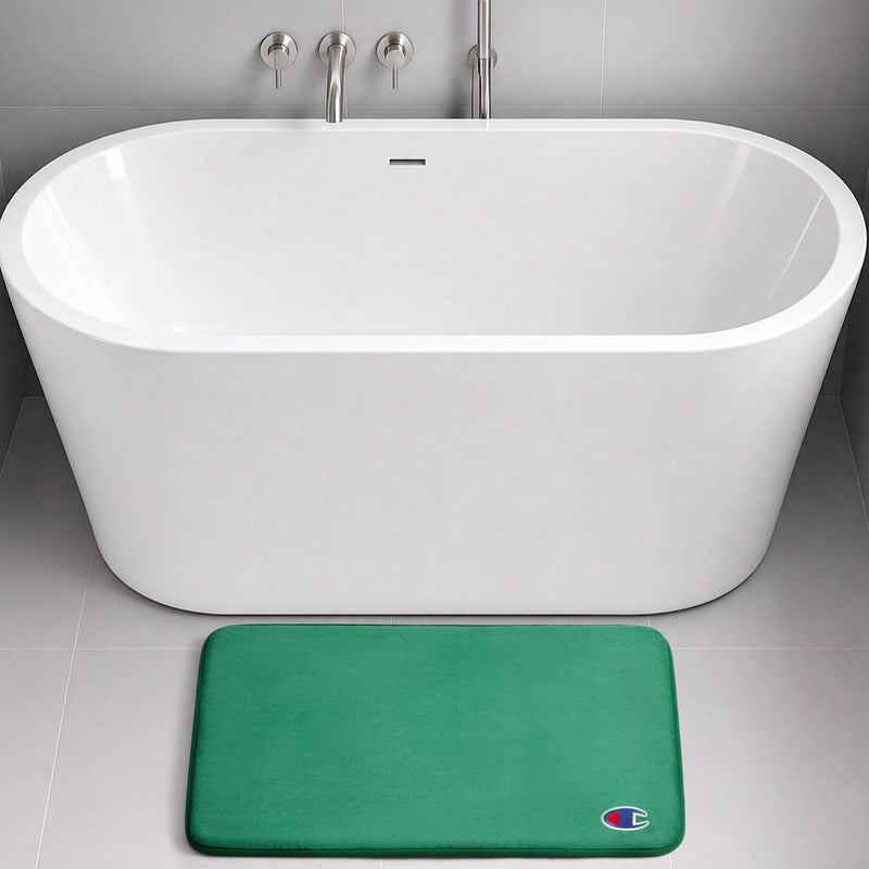 A modern white bathtub with wall-mounted fixtures sits on a gray tile floor. In front of the tub is a green Logo Patch Memory Foam Bath Mat from Champion, featuring a small, colorful C logo in the bottom right corner.