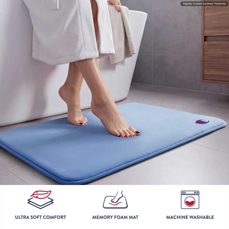 A person steps out of the bathtub onto a light blue Champion Logo Patch Memory Foam Bath Mat in a modern bathroom. Below, icons and text highlight its plush comfort, luxurious memory foam material, and convenient machine washability.