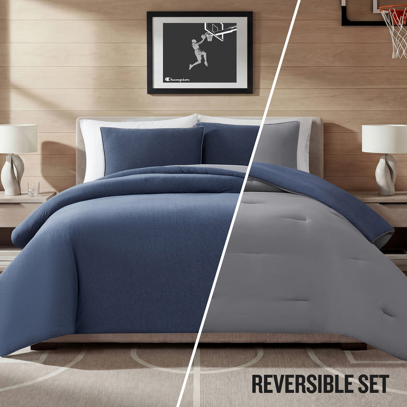 Transform your space with the Champion Reversible Solid Jersey Comforter Set, showcasing an athletic-inspired bedding look. The comforter features a split design—one side blue, the other gray—and is paired with matching lamps, nightstands, and a framed basketball art piece above the bed. Text reads: REVERSIBLE SET.