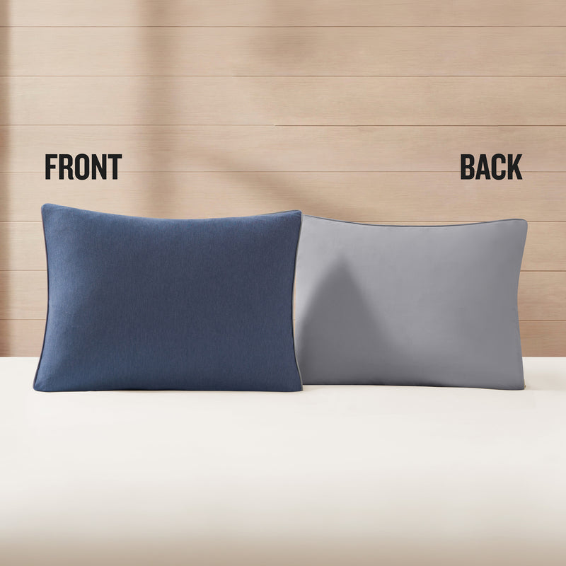 Two pillows from the Champion Reversible Solid Jersey Comforter Set are placed side by side on a bed against a wooden wall; the left pillow displays the dark blue front while the right pillow reveals the light gray back, emphasizing the reversible design with FRONT and BACK labels above each pillow.