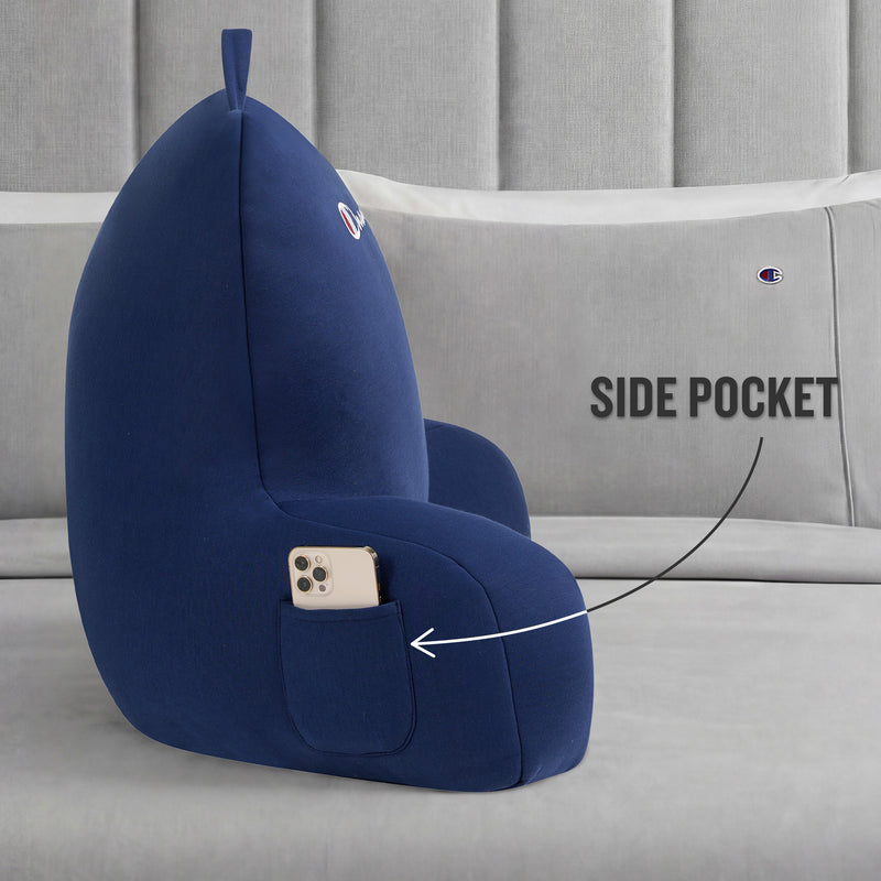 A Champion Jersey Backrest Pillow in blue with armrests rests on a bed, made from soft jersey knit fabric. An arrow highlights the convenient side pocket that holds a smartphone. Both the pillow and bedding display a small embroidered Champion logo.