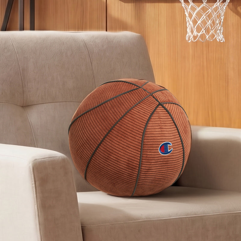 A Champion Sport Shaped Pillow in the style of a large basketball rests on a beige armchair, adding a playful corduroy accent to the scene. In the background, there’s a wooden wall with a basketball hoop mounted above.