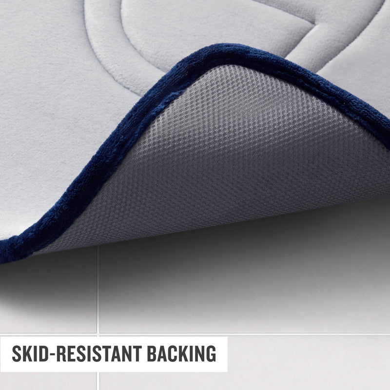Close-up of a gray Champion Quilted Logo Memory Foam Bath Mat with a dark blue border slightly lifted, revealing its textured, skid-resistant backing on a tiled floor. Text in the corner reads, SKID-RESISTANT BACKING.