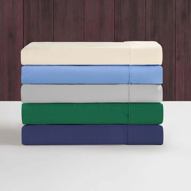 A neatly stacked set of five folded Champion Elastic Logo Microfiber Sheet Sets, in cream, light gray, light blue, green, and navy blue, rests on a white surface against a dark wood-paneled background.