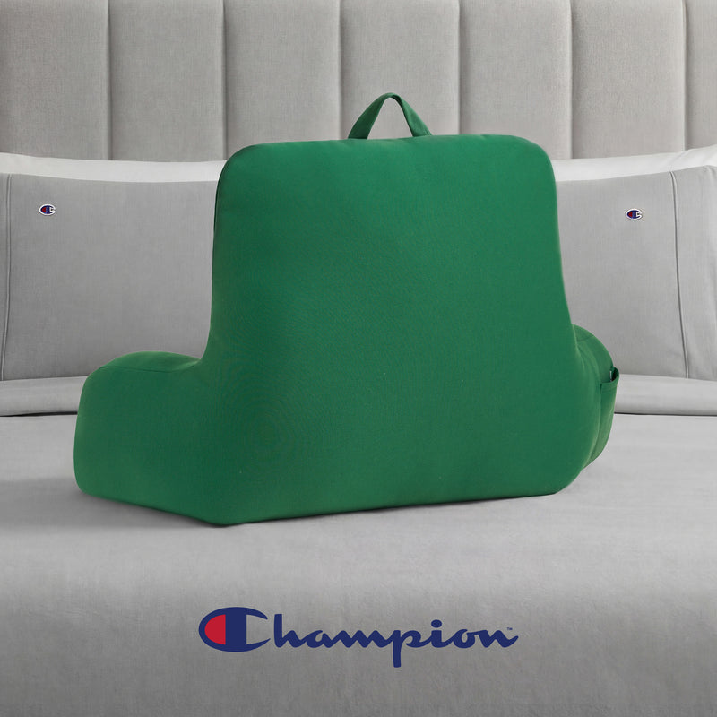 A Champion Jersey Backrest Pillow in green, featuring a convenient handle, rests on a bed with light gray Champion bedding. The iconic Champion logo is visible on the bed next to the backrest pillow, lending a sporty vibe to the cozy scene.