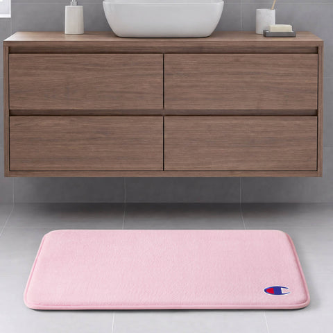 A light pink Champion Logo Patch Memory Foam Bath Mat featuring a small logo in the corner is placed on a gray tile floor in front of a floating wooden bathroom vanity with drawers. This skid-resistant memory foam mat combines style and safety for your space.