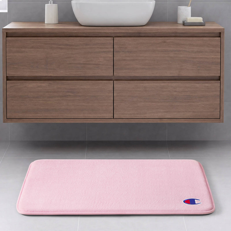 A light pink Champion Logo Patch Memory Foam Bath Mat featuring a small logo in the corner is placed on a gray tile floor in front of a floating wooden bathroom vanity with drawers. This skid-resistant memory foam mat combines style and safety for your space.
