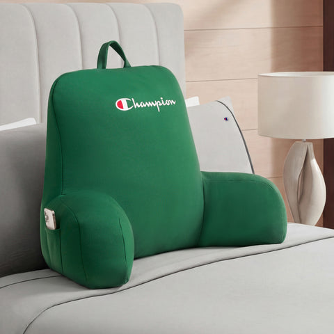 A large green Champion Jersey Backrest Pillow with supportive armrests and “Champion” embroidered on it sits on a neatly made bed in a modern bedroom.
