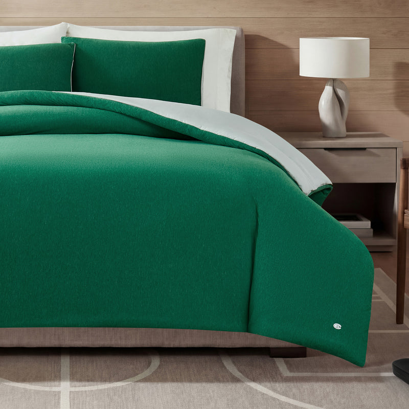 A bed featuring the Champion Reversible Solid Jersey Comforter Set in green with a matching pillow, complemented by white sheets and accompanied by a bedside table with a white lamp, all set against a wood-paneled wall and atop a beige rug with a curved pattern.