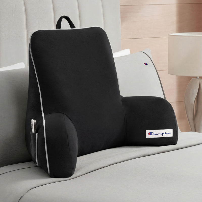 A Champion Faux Wool Backrest Pillow with supportive arms and handy side pockets, distinguished by the brand’s logo, rests on a neatly made bed dressed in light gray linens and accented by a tufted headboard. Its plush faux wool cover brings added comfort and style to the space.