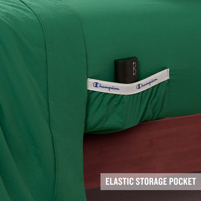 A green Peak Comfort Microfiber Sheet Set by Champion, featuring an elastic storage pocket labeled “Champion” that holds a small black item. Designed for both performance and comfort, this athletic bedding set includes deep pocket sheets from the Champion collection. The bed frame is wooden, with the text “ELASTIC STORAGE POCKET” displayed in the bottom right corner.