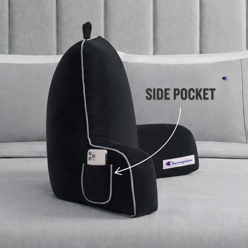 A black Champion Faux Wool Backrest Pillow with supportive arms rests on a bed. An arrow highlights the side pocket containing a smartphone, with text that reads, SIDE POCKET.