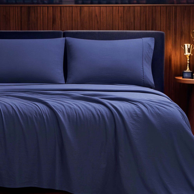 A neatly made bed features the Champion Elastic Logo Microfiber Sheet Set in navy blue, complete with matching pillowcases, set against a wooden headboard. On the small round nightstand sits a gold trophy-shaped lamp.