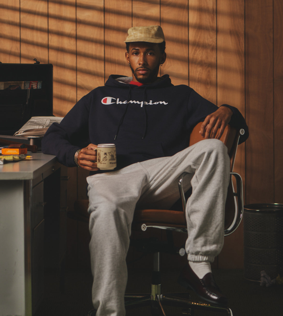 Man sitting in a chair holding a mug, wearing a Champion hoodie and light gray pants, in a room with wooden walls.