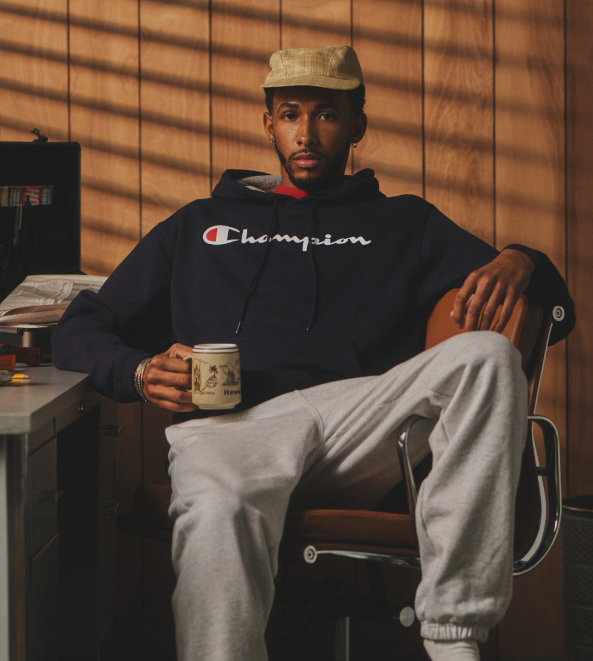 Person wearing a Champion hoodie sitting in a chair with a mug, against a wooden paneled wall.