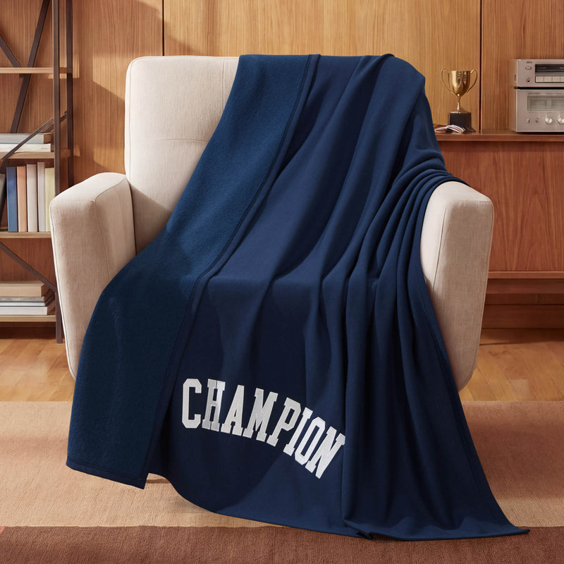 A Jersey with Fleece Backing Throw Blanket by Champion, featuring bold white CHAMPION lettering on navy blue, is draped over a beige armchair in a wood-paneled room—capturing the spirit of athletic bedding—with a trophy and stereo displayed on a cabinet in the background.