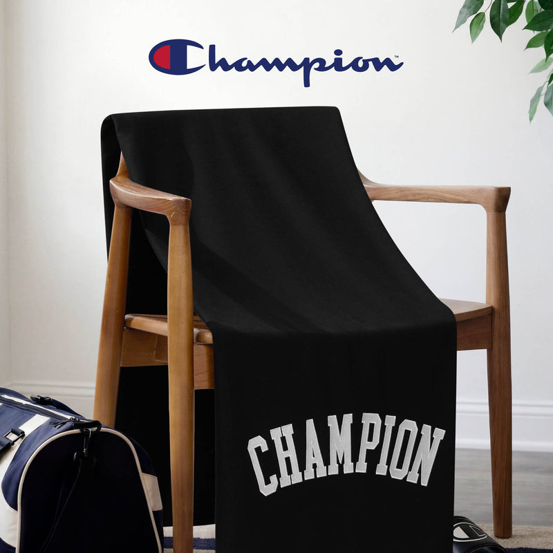 A Champion Jersey with Fleece Backing Throw Blanket in black is draped over a wooden chair. The word “CHAMPION” appears in bold white block letters along the bottom edge, with the signature Champion logo displayed at the top, emphasizing its iconic design. Nearby on the floor sits a blue and white duffel bag.