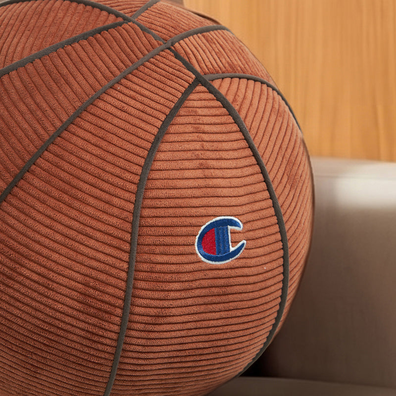 A Champion Sport Shaped Pillow featuring a cozy corduroy basketball design rests on a beige couch, adding a sporty accent to the space in front of a wooden wall background.