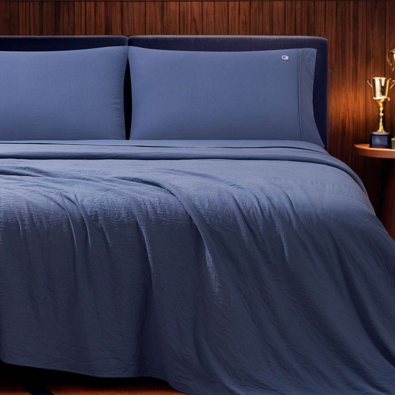 A neatly made bed features the Champion Embroidery Patch Microfiber Sheet Set in blue, paired with matching pillows and set against a dark headboard and wood-paneled wall. Beside the bed, a small round table topped with a gold trophy completes the winning look.