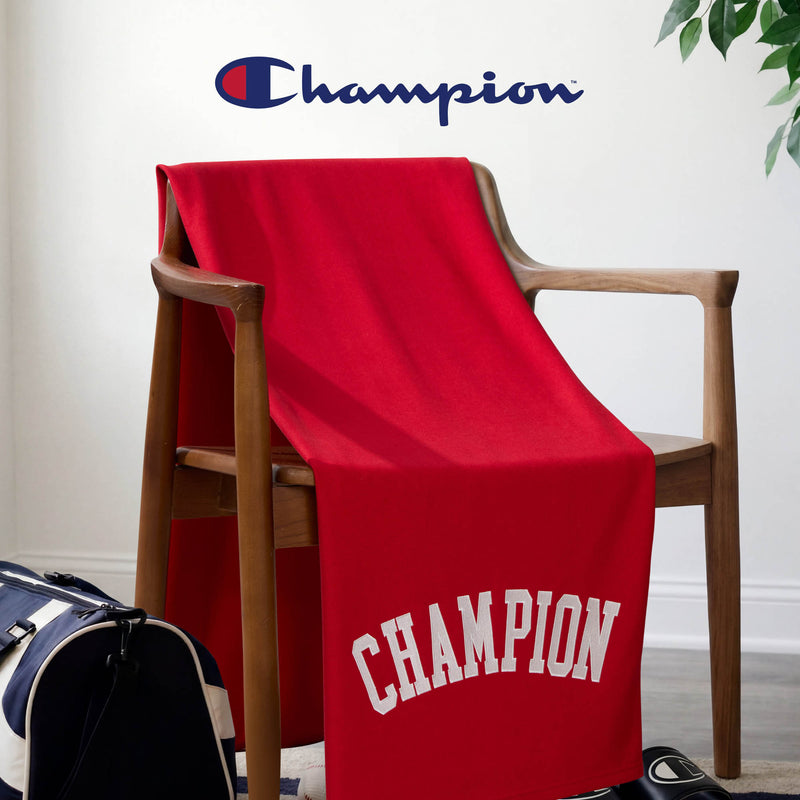 A Champion Jersey with Fleece Backing Throw Blanket in red, featuring “CHAMPION” in bold white letters, is draped over a wooden chair. The iconic Champion logo is displayed on the wall above, while a navy and white gym bag sits on the floor nearby, creating an athletic bedding vibe.