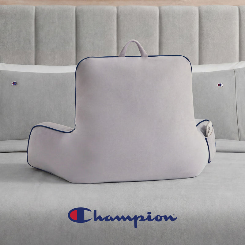 A Champion Faux Wool Backrest Pillow in light gray, featuring supportive arms, a handle, and a side pocket, is placed on a gray bed. The bedspread displays the Champion logo in blue and red, and matching Champion-branded pillows complete the bedding ensemble.