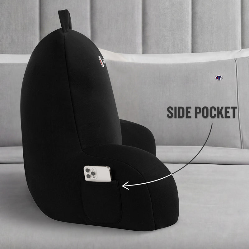 A Champion Jersey Backrest Pillow in black is displayed on a bed with gray pillows, showcasing its iconic logo and a handy side pocket holding a smartphone. An arrow points to the pocket with the label “SIDE POCKET,” emphasizing this pillow’s functional design.