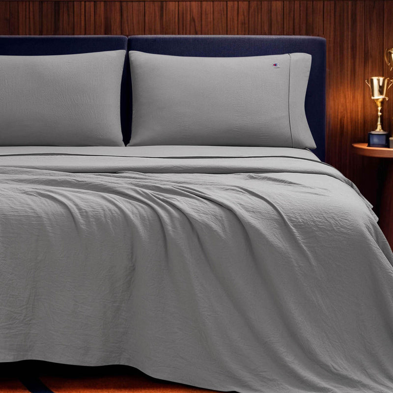 A neatly made bed with light gray Champion Embroidery Patch Microfiber Sheet Set and two pillows, set against a dark upholstered headboard and a wood-paneled wall. A small trophy sits on a nightstand to the right.
