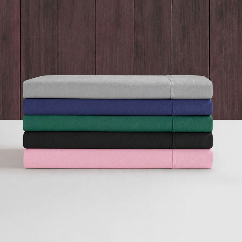 A neatly stacked pile of five folded Peak Comfort Microfiber Sheet Sets by Champion in gray, blue, green, black, and pink—each set featuring deep pocket sheets—is displayed against a dark wooden wall and white surface.