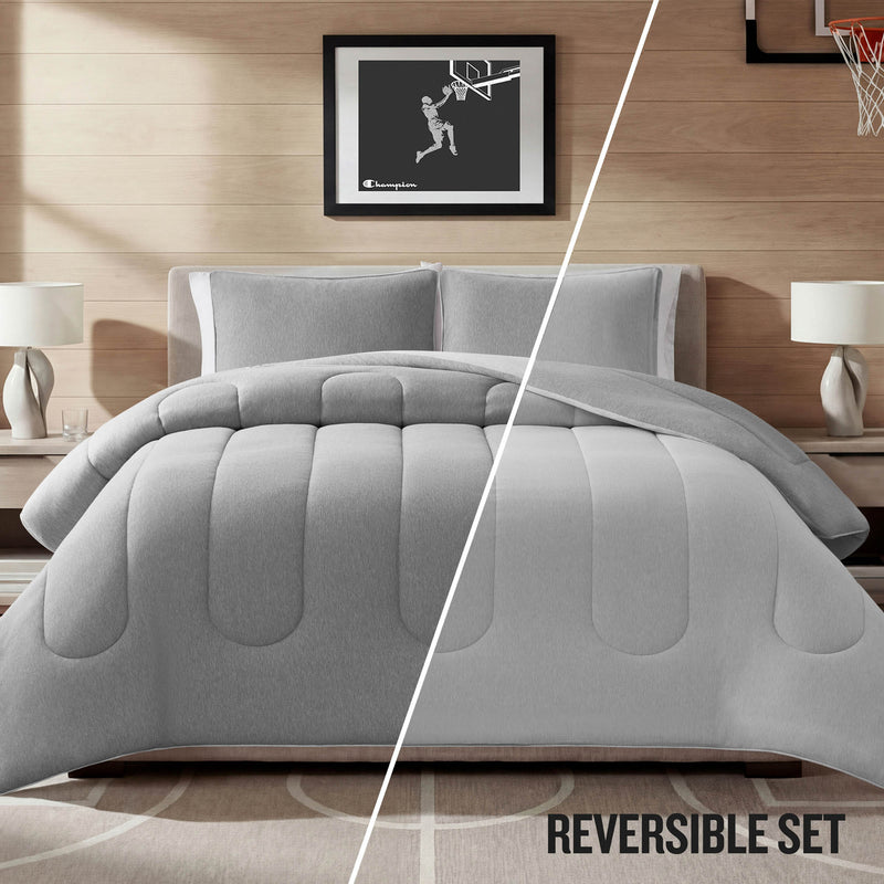 A Champion Premium Reversible Solid Jersey Comforter Set in gray is displayed on the bed, with the comforter split to reveal both reversible sides of its jersey design. The room includes two lamps, nightstands, and a framed basketball picture on a wooden wall. Text reads: REVERSIBLE SET.