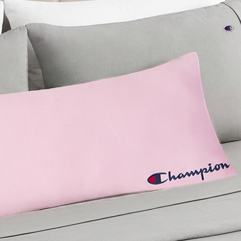 A Champion Jersey Body Pillow in light pink, featuring the iconic blue and red Champion logo, rests on a grey bed alongside pillows in grey and white covers—including another pillow embroidered with the Champion logo—bringing an athletic accent to the room.