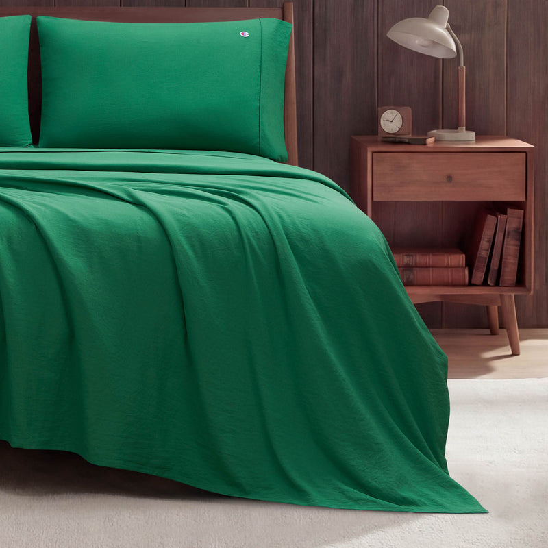 A neatly made bed features bright green Champion Peak Comfort Microfiber Sheet Set and matching pillowcases, positioned beside a wooden nightstand topped with a lamp, books, and a small clock, all set against a dark wood paneled wall.