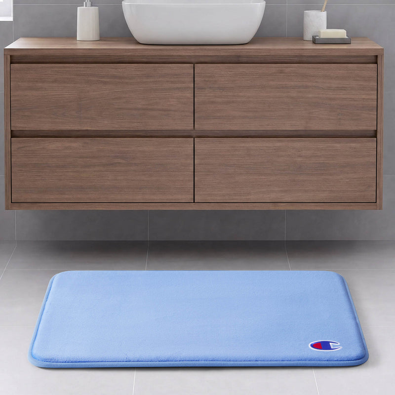 The Champion Logo Patch Memory Foam Bath Mat in light blue features a rectangular design with a small logo patch in the corner. Crafted from soft memory foam for enhanced comfort and skid-resistant backing for safety, this mat sits elegantly on a grey tiled floor in front of a wooden bathroom vanity with four drawers and a white sink, bringing style and practicality to your space.