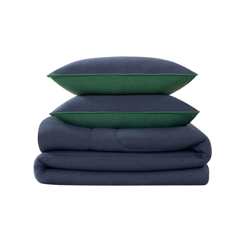 Two dark blue pillows with green edges from the Champion Premium Reversible Solid Jersey Comforter Set are stacked on top of a neatly folded matching comforter, all placed against a plain white background.